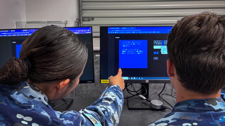 A teenage girl and boy in AAFC uniform sit facing two computer screens